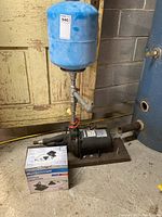 Jet pump with blue pressure tank and boxed utility pump on floor in basement.