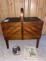 Front view of wooden sewing cabinet with handle and closed hinged drawers. Metal container with sewing notions and Valley-Bee Metal Weaving Loom box placed in front.