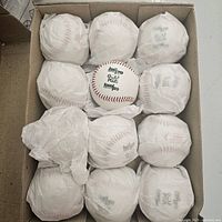 Top-down view of 12 baseballs arranged in cardboard box with one ball fully visible and other balls wrapped in tissue paper.