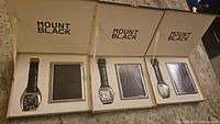 Three Mount Black watch and money clip sets in presentation boxes on countertop.