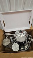 Box containing shiny stainless steel pots with matching lids and stainless steel handles alongside lidded glass jars, all neatly packed.