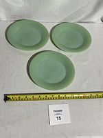 Three pale green translucent Fire-King Jadeite salad plates arranged on white cloth surface with measuring tape showing approx 6.5 inch diameter.