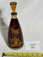 Front view of amber glass decanter bottle with an etched bird and reeds design, alongside measuring tape showing approx. 10 inches in height.