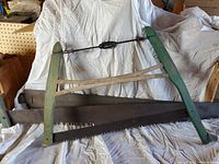 Three vintage crosscut saws displayed against a white backdrop, one with a green wooden two-man frame and tensioning mechanism, and two straight steel blades with saw teeth.