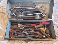 Open metal tool box showing various hand tools including adjustable wrenches, chisels with wooden handles, and files.