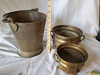 Full view of copper ash pail with handle and two brass planters with handles and feet, placed next to a ruler for scale.