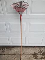 Full view of metal rake with wooden handle and red metal tines leaning against garage door