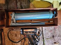 Blue metal HP multi load grease gun with lever handle inside cardboard box on worn wood surface, viewed from above.