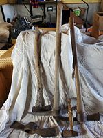 Full view of four pick axes standing upright against a white cloth background in a cluttered room. Wooden handles are worn and metal heads heavily rusted indicating usage and age.