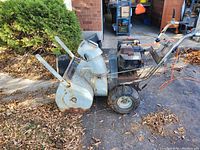 Side view of the snowblower showing rusted metal body, chute, and wheels with rubber tires.