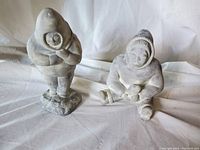 Both soapstone Inuit figurines together on a white cloth background, one standing and one sitting.