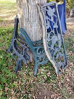 Multiple wrought iron bench arms and a blue wrought iron back panel featuring floral motifs leaning against a tree outdoors on grass and leaves.