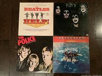 Four vinyl LP records displayed on wood flooring. Albums shown from front covers: The Beatles 'Help!' soundtrack, KISS, The Police 'Outlandos d'Amour', and Aerosmith.
