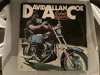 Front cover of David Allan Coe 'Rides Again' LP showing the artist riding a motorcycle with visible wear and minor record scratches mentioned by seller.