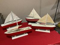 Four handcrafted sail boats painted red, white, and black on white stands displayed on red cloth.