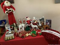 Full view of Christmas holiday items arranged on a red tablecloth including several Santa Claus figurines, deer and reindeer, ornaments, nutcrackers, mugs, and decorations