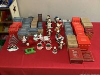 Photo showing multiple boxed Avon vintage Christmas ornaments and several out-of-box ornaments including figurines of Santa Claus, snowmen, reindeer, and cows arranged on a red cloth.