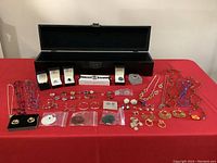 Full view of costume jewelry including necklaces, rings, earrings laid out on a red cloth with black wooden box behind.