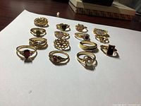 Fifteen gold-colored ladies rings arranged on white paper showing various styles and stones.
