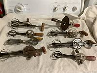 Eight vintage metal hand beaters with wooden handles arranged on white fabric, showing various designs and wear.