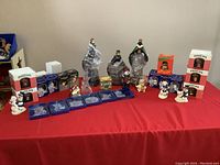 Overview of lot showing large Wisemen figurines, numerous boxed and unboxed ornaments on red tablecloth.