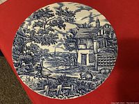 Blue and white decorative plate with detailed pastoral scene including figures, buildings, trees, and animals.