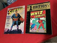 Two comic books on red cloth: 1974 Superman Limited Collectors Edition and 1940 Whiz Comics 1st Edition showing their front covers.