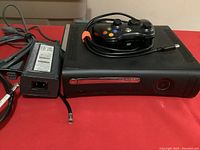 Xbox 360 Elite console with power supply and wired controller on top, all laid on a red surface.