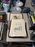 Top view of stacked art prints, some framed with wooden frames, showing botanical themes and pineapple plant illustration visible.