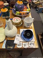 Photo of assorted ceramic ashtrays, coasters, mugs, decorative tile, and white embossed bottle arranged on wooden table