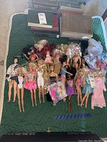 Overall view of collection with approximately 19 dolls, some fully dressed, some partially dressed or missing attire, with doll accessories in plastic bags. Dolls arranged on green quilted surface alongside some books.