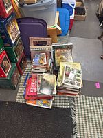 Photo showing various hardcover and softcover books stacked on a table with visible titles in gardening, dictionary, history, and bird-themed books. A pair of carved wooden bird bookends visible supporting some books.