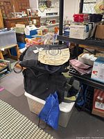 View of several carry bags including a beige tote bag with Georgian Court College print on top of black bags and a small blue drawstring bag in front, placed on storage bins and shelf in a room.