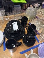 Black colored china set including stacked dinner plates, bread plates, bowls, and several mugs with handles shown on a table with other glassware around them.