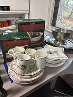 Overall display of boxed Pfaltzgraf Snow Village serving bowl and sugar/creamer set alongside Johnson Brothers Christmas-themed plates, mugs, and bowls with visible Christmas motifs.