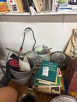 Photo showing stacked metal mixing bowls with kitchen utensils inside, silverware in green ceramic bowl, assortment of cookbooks stacked alongside, and clear Pyrex glass casserole dish with lid.