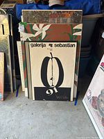 Nine framed art pieces stacked and leaning against each other showing front details of the two most visible art pieces - one a brown floral print and the other a black and white graphic print with text