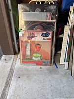 Five stacked framed posters leaning against a wall, sealed in plastic wrapping.