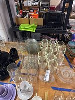 Variety of glassware including clear tumblers, whiskey glasses, shot glasses, and bowls on a table, black and green accessories visible in background.