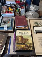 Overview of an assortment of books including history and art books and framed botanical prints, placed on a table with other household items in the background.