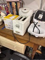 Photo showing white two-slice toaster, white Toastess sandwich maker with black strap, and boxed Dash Rapid Egg Cooker on wooden surface with other kitchen items.