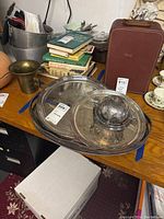Set of silver plated English serving plates and tray with dome cover shown on wooden table with additional items and vintage suitcase nearby.