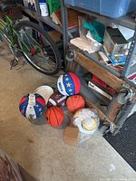 Photo shows two red, white, and blue basketballs with star pattern and Ultra branding, two orange basketballs, one brown and white football, a yellow and white soccer ball, and a stack of white frisbees on garage floor near shelving and a bicycle.
