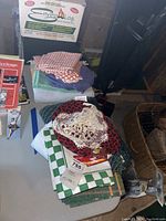 Table linens including checkered napkins, crocheted doilies, various cloths stacked on a table, with a box and miscellaneous items in background.