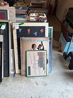 Stack of framed posters and wall art leaning against a wall, showing visible wear and different sizes.