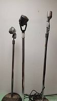 Three antique microphones on metal stands with round bases against a plain wall.