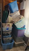 Photo showing stacked plastic storage bins and totes of various sizes and colors leaning against a wall near a window.