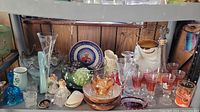Photo showing assortment of glass vases, bowls, stemware, figurines including frosted angels, decorative plate with portrait, and various small glass and ceramic items