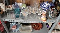 Wide view on gray shelf of assorted glassware and ceramics including Murano-style vase, stemware, blue/white tureen and plates, small glass jars, and figurines.