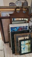 Photo showing a wooden framed mirror with carved decorative top alongside multiple framed artworks leaning on the floor.